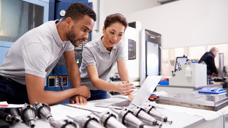 Two engineers - one man and one woman from different races are checking a laptop screen with equipment in front of them on the table -doing resource scheduling
