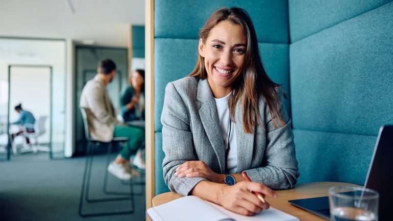 business woman in her late 20s looking at the camera smiling, in a modern cozy office